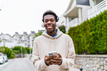 Young African man wearing headphones, smiling and holding a smartphone in a city street.