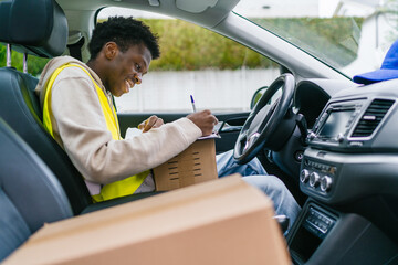 Young African courier writing on a clipboard inside vehicle, preparing package delivery