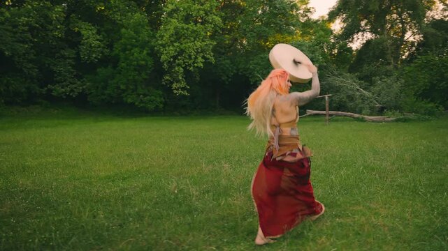 Sexy fantasy happy woman shaman holds drum in hands back rear view, plays music beats melody runing dancing. Magic forest green grass trees summer nature. Fairy Girl boho style dress red hair art 4k