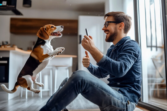 A man joyfully interacts with his jumping beagle dog indoors during a sunny afternoon at home, capturing the moment with his smartphone