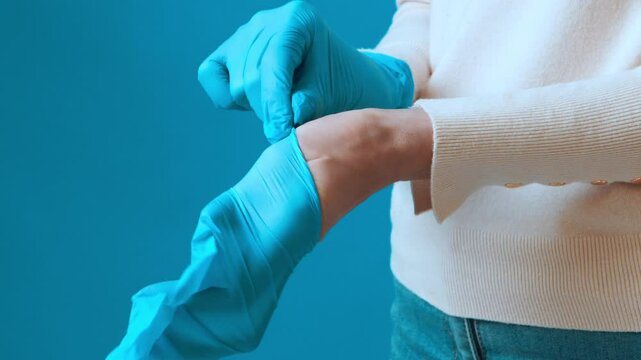 A person is wearing a light-colored sweater while putting on blue disposable gloves. The setting features a bright blue backdrop, creating an engaging visual contrast.