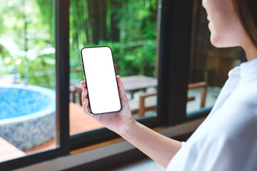 Mockup image of a woman holding mobile phone with blank desktop white screen at home