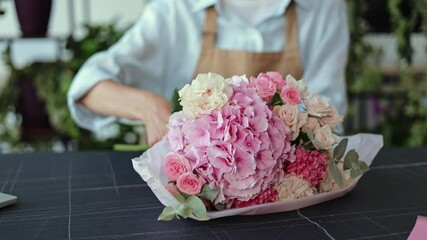 A florist is working on a flower arrangement for a delivery order in a flower shop. She is cutting stems and arranging pink and white roses and other flowers in a bouquet.