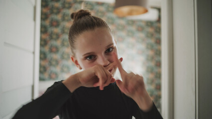 Adorable and happy young girl with make up and bun hairstyle makes funny hand gestures on camera. Cheerful Caucasian teenager smiles and looks at camera while having fun at home. Lifestyle concept.