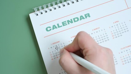 Female hand circling date on calendar on green background, top view.