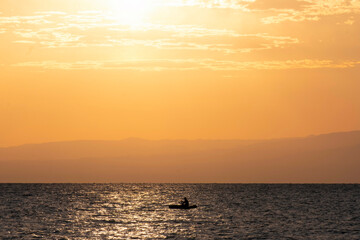 Silhouette of a fisherman on a boat in the sea or lake against the backdrop of water and a sunrise or sunset.