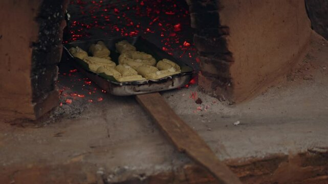 Man placing a baking tray with chipas and helping a woman put it inside the tatakua with coals inside. The tatakua is a traditional Paraguayan clay oven, commonly used to make chipa during Holy Week