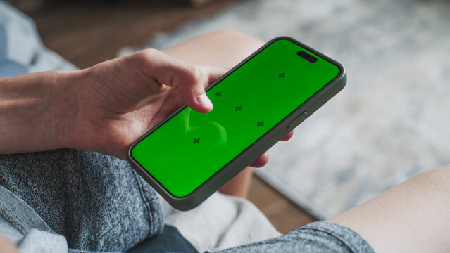 Close up of teenager using mobile phone with green screen, surfing Internet or scrolling social media while sitting on sofa and spending time at home. Chroma key displayed on smartphone screen. Mockup - Powered by Adobe