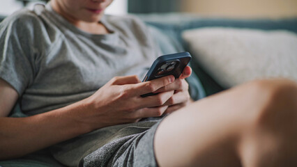 Close up of Caucasian teenager chatting with friends or scrolling content in social network using mobile phone while sitting on sofa. Young boy resting and spending weekend at home. Lifestyle concept.