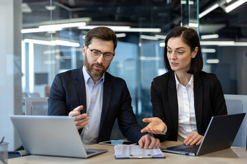 Two business professionals engage in discussion during a meeting, using laptops and documents...