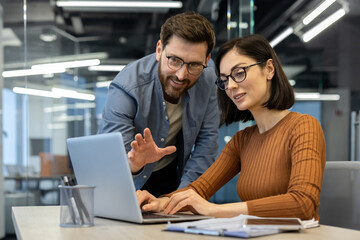Two colleagues collaborating in office, focused on project. Man pointing at laptop screen, woman typing. Teamwork, technology, and planning concepts. Pair engaged with office work