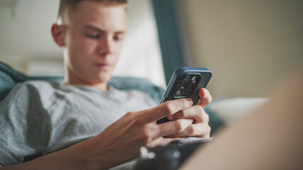 Caucasian teenager chats in messaging app with friends or watches content using mobile phone while sitting on sofa in living room. Teen boy spending weekend at home. Lifestyle concept. Close Up.