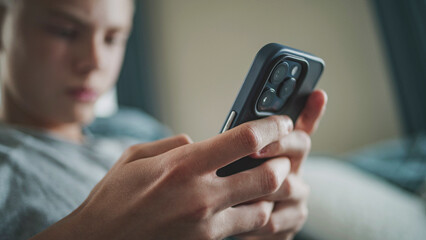 Close up of teenage boy typing and chatting with friends in messaging application using mobile phone while sitting on sofa. Caucasian teenager relaxing and spending weekend at home. Lifestyle concept.