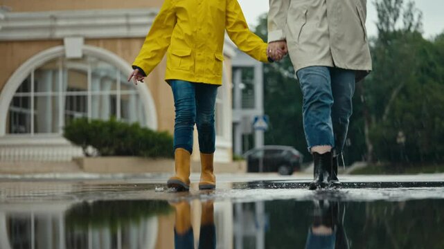 happy girl in a yellow jacket and rubber boots walks through a puddle with her mother and splashes water to the sides in the park after the rain