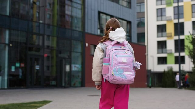Young girl, around 7-9 years old, is seen from behind as she walks toward school building, wearing grey varsity jacket, pink pants, carrying pink backpack. School ahead is modern, with glass windows.