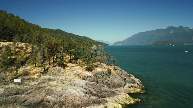 Drone shot flying around a rocky peninsula located on Bowen Island surrounded by trees, mountains, cliff edges and clear blue tropical water for swimming on a sunny day in Vancouver