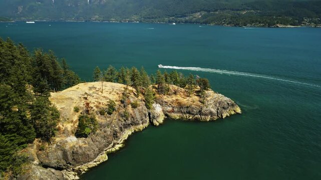 drone shot as speedboat sails around the small peninsula of a small tropical island covered in sandy rocks and trees surrounded by clear blue water on a sunny day