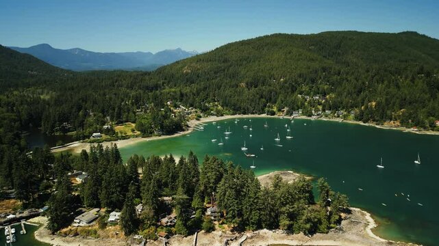 Slow drone orbit around a tropical bay port on Bowen Island near Vancouver with boats anchored in the clear blue water surrounded by rolling hills covered in trees on a sunny day