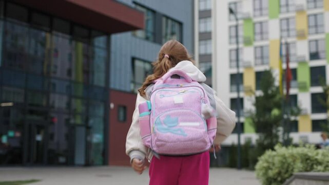 Young girl, around 7-9 years old, is running towards the school building. She is wearing a grey and white varsity jacket, bright pink pants, and a large pink backpack with blue accents.