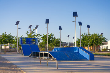 Skate park with ramps and rails in a clean urban environment. The area is lit by several solar-powered street lights, highlighting the sustainable energy infrastructure. Spain, Europe