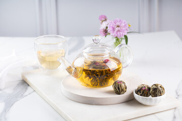 Tea in a glass teapot with a blooming large flower. Teapot with exotic green tea-balls blooms flower on light grey background. Tea ceremony.