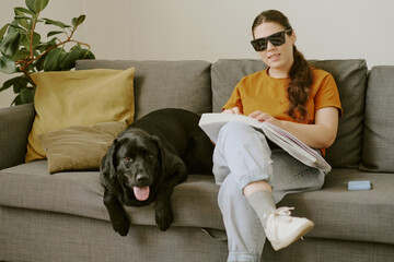 Young woman with disability sitting on sofa and reading Braille book while her pet lying next to her
