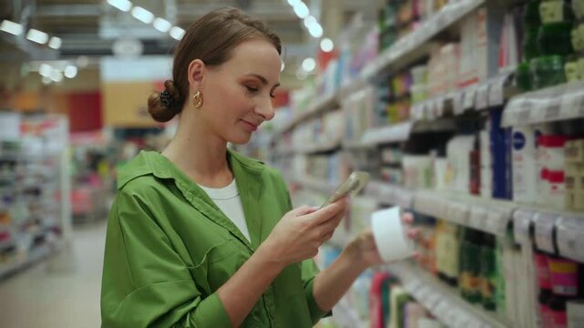 Customer compares prices and reads product information on her smartphone while shopping for beauty items in a grocery store aisle filled with cosmetics and skincare products