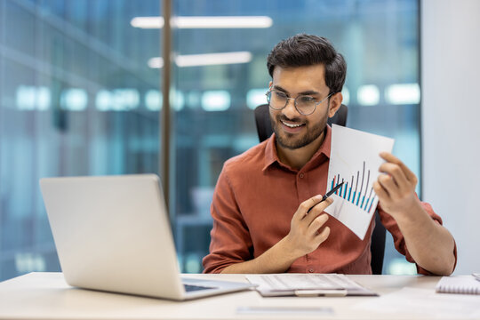 Businessman engaging in video call holding growth chart, analyzing data at office. Professional sitting at desk with laptop, focusing on presentation and communication in contemporary workplace.