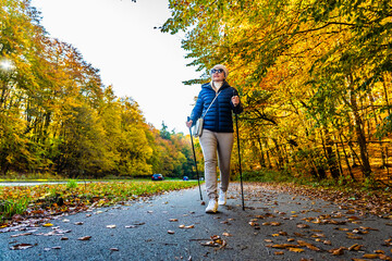 Beautiful middle-aged blonde woman in casual wear walking with poles on sidewalk by street in...