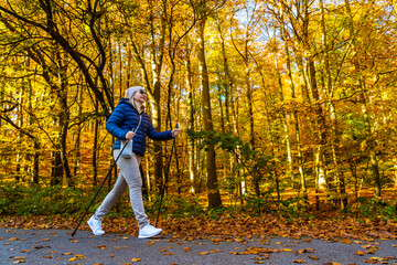 Fototapeta premium Beautiful middle-aged blonde woman in casual wear practicing Noridc walking on sidewalk in autumn scenery. Side view. Autumnal Nordic walking in city.
