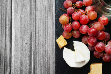 A bunch of large pink grapes next to Brie and Parmesan cheese on a black tray on a gray wooden background