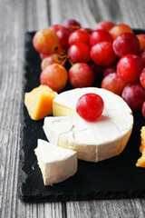 A bunch of large pink grapes next to Brie and Parmesan cheese on a black tray on a gray wooden background