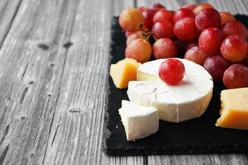 A bunch of large pink grapes next to Brie and Parmesan cheese on a black tray on a gray wooden background