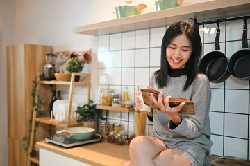 Pleased young woman drinking milk and reading book while sitting on kitchen counter