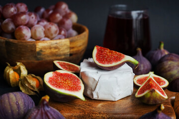 Ripe whole and sliced ​​figs with brie cheese, Physalis fruits, pink grapes on a wooden board and a glass of red wine on a dark background