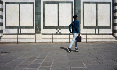 A young man walks briskly with a bag against a striking marble wall backdrop. Ideal for themes of travel, urban life, and dynamic movement in modern city settings.