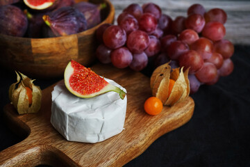 Ripe figs next to Brie cheese on a wooden board, pink grapes and Physalis fruits on a dark background