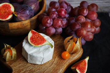 Ripe figs next to Brie cheese on a wooden board, pink grapes and Physalis fruits on a dark background