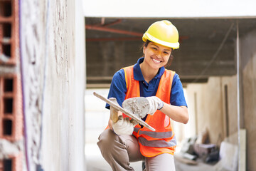 Portrait of smiling young female bricklayer taking cement on trowel from bucket while plastering...