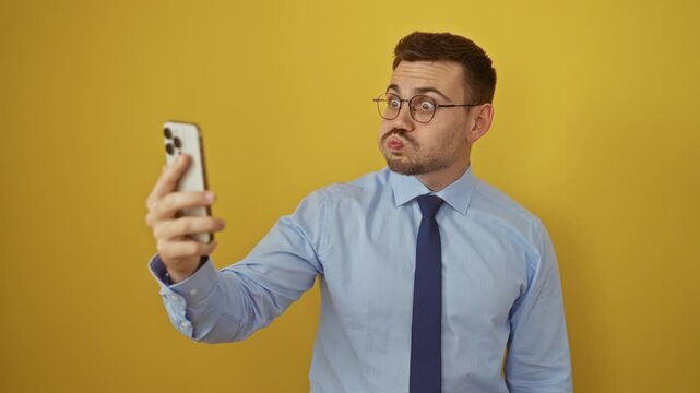 Young hispanic man in shirt and tie making a fish face with crazy eyes and mouth while using smartphone, over isolated yellow background, comical and silly expression