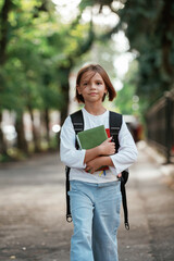 Walking forward and holding notepad. Schoolgirl with backpack is outdoors