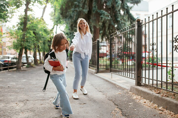 Daughter is holding the woman's hand and leading the way. Schoolgirl with her mother are outdoors...