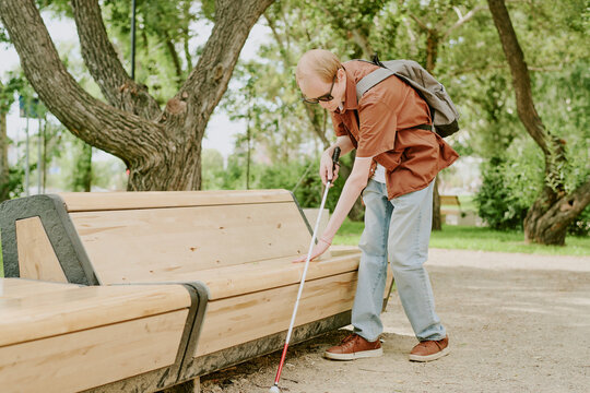 Caucasian blind man using metal cane to walk in park alone, he touching bench