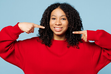 Little happy kid teen girl of African American ethnicity wear red sweatshirt point fingers on toothy smile white teeth isolated on pastel plain light blue cyan background. Childhood lifestyle concept.