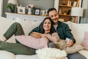 Diverse couple, watching tv and remote on living room couch with smile, hug or relax with click for...