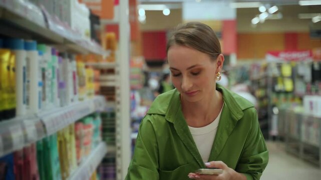 Customer wearing casual clothing is comparing products while shopping in a grocery store. She is using her phone to scan barcodes and compare prices