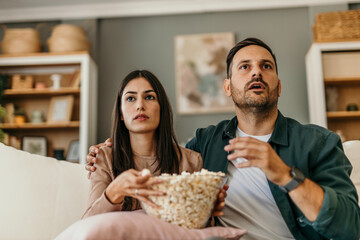 Multiracial husband and wife eating popcorn from a bowl and watching drama movie while chilling on the couch in weekend