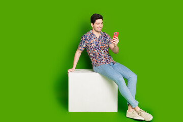 Full size photo of positive guy dressed print shirt jeans sit on white platform look at smartphone isolated on green color background