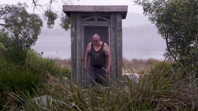 A man dressed in casual attire stands outside a quaint wooden toilet, holding a newspaper under his arm. The atmosphere is calm and slightly overcast, surrounded by greenery and a lake.