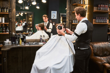 Experienced hairdresser styling client's hair. Handsome hairstylist in fancy outfit at work. Male...